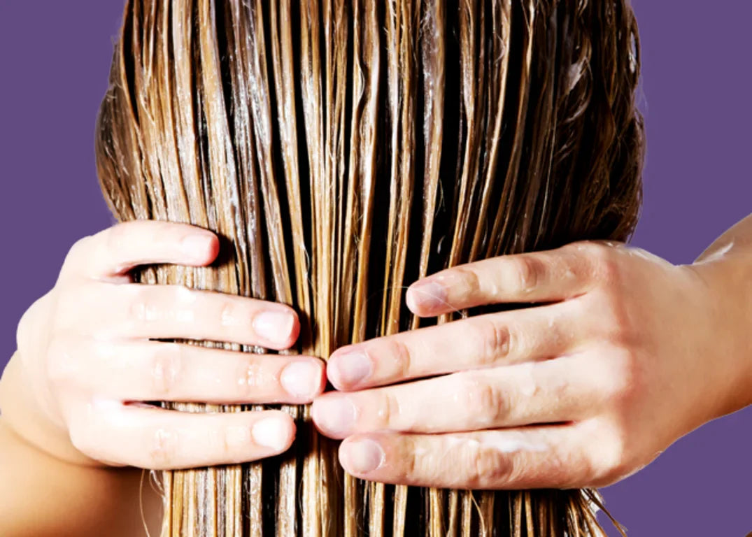 A woman with long and wet, brownish blonde hair holds it behind her head with both hands as she detoxes it.
