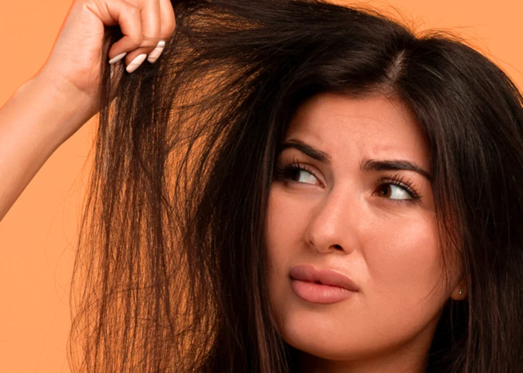 Tan woman with straight, dark and damaged hair looks upset at it while holding it up in her hand