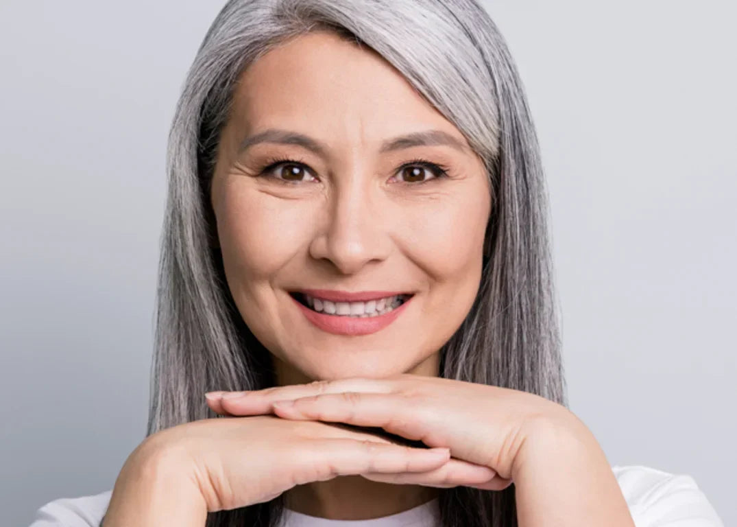 A woman with long gray hair smiles facing forward with her hands below her chin in front of a white background.