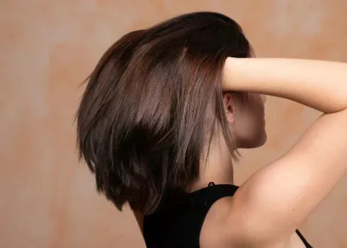 A fair-skinned woman’s hands touch her brown hair to show her balanced and dandruff-free oily scalp in front of an orange background.