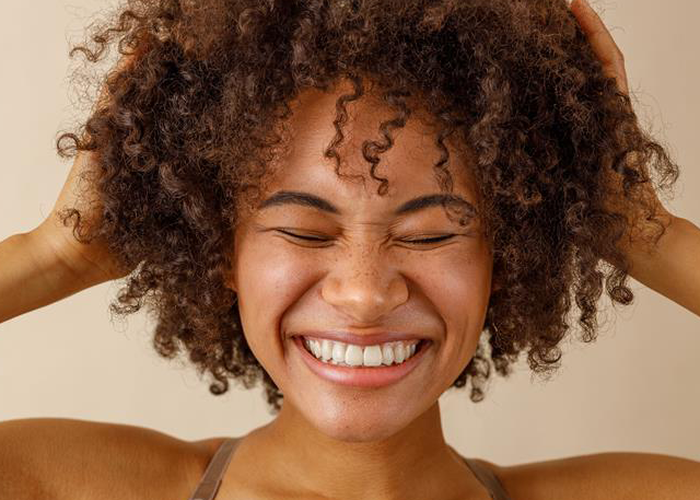 Cheerful tan-skinned woman with voluminous shiny curly brown hair on beige background demonstrating healthy hair cuticles