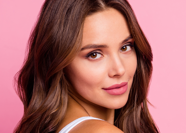 A woman with brown wavy hair and tan skin looks back in front of a pink background.