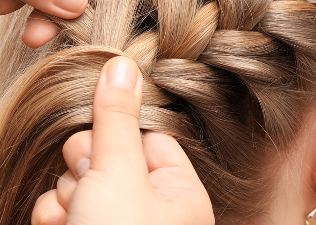 A fair-skinned woman with blonde hair is having her hair braided for a festival hairstyle in front of a white background.