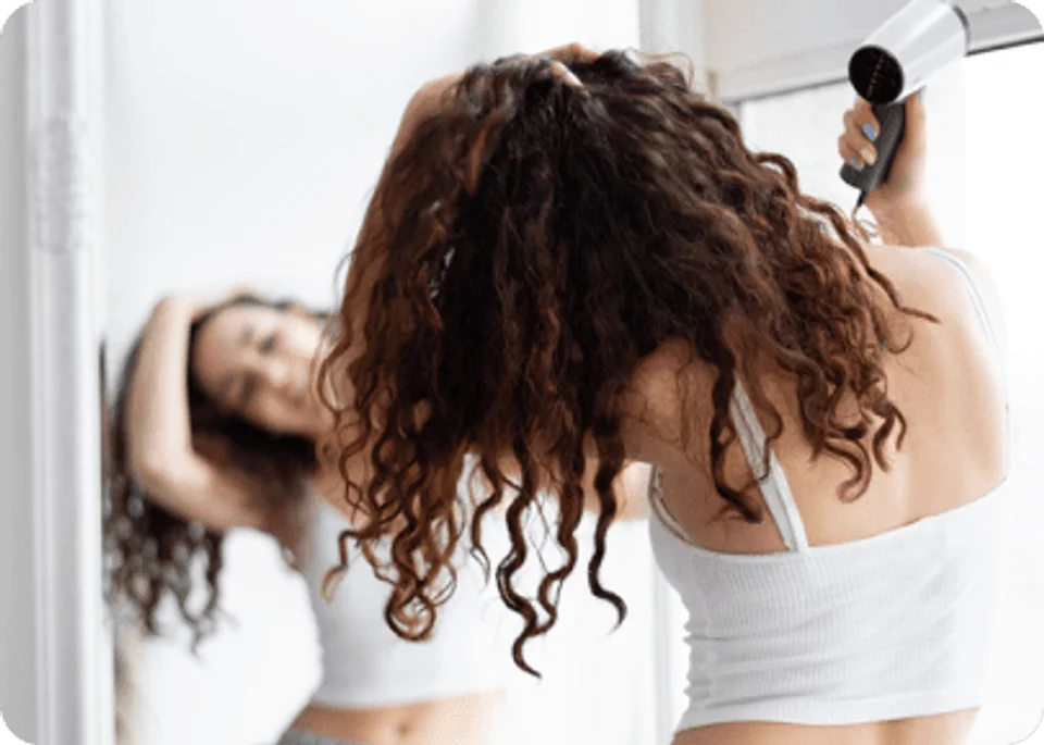 Young brunette girl female woman adult in grey shorts and white top drying her curly hair with blow dryer after shower in front of the mirror.