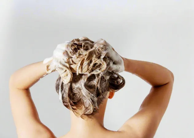 A Fair skinned woman with blonde hair uses dandruff shampoo to wash her hair in front of a grey background.