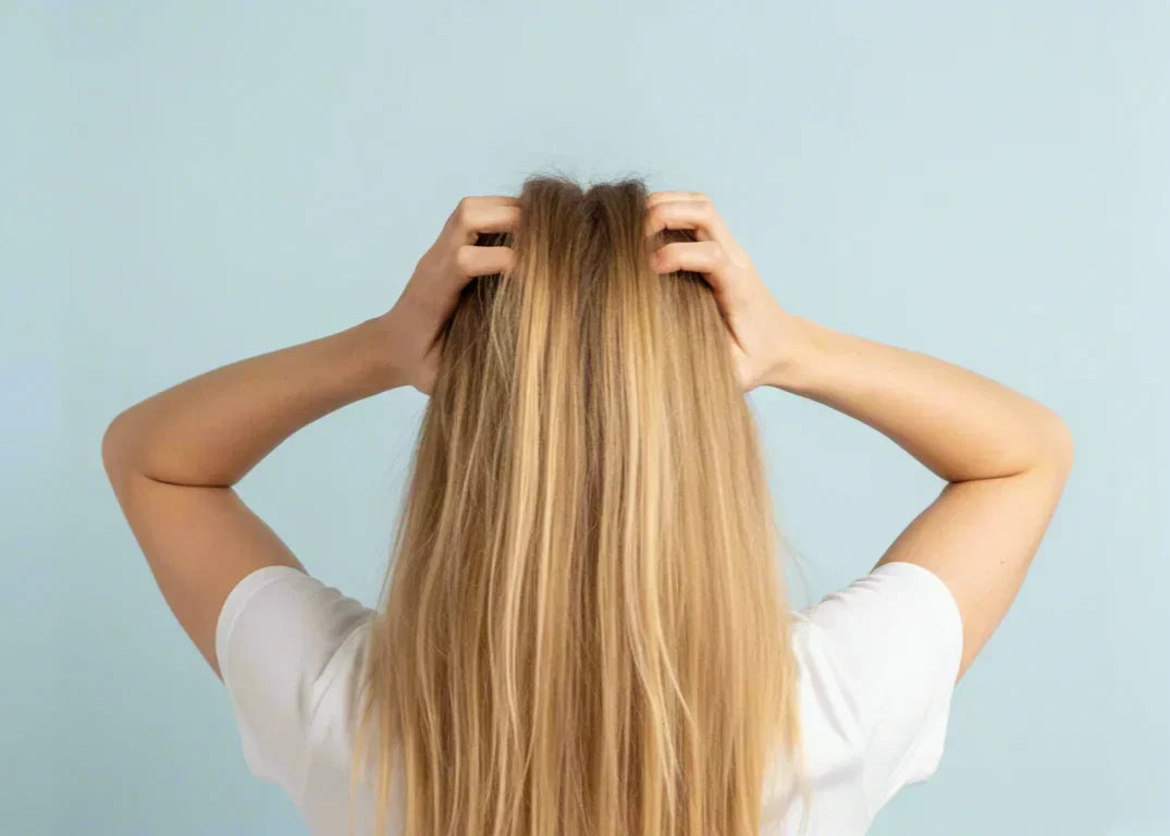 A tan-skinned woman scratches her blonde hair due to her itchy and flaky scalp in front of a blue background.