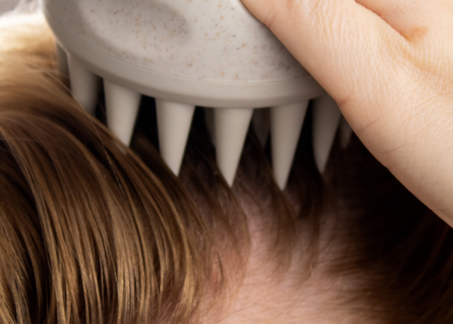 A fair-skinned hand uses a grey scalp massager to massage through hair in front of a brown-grey background.