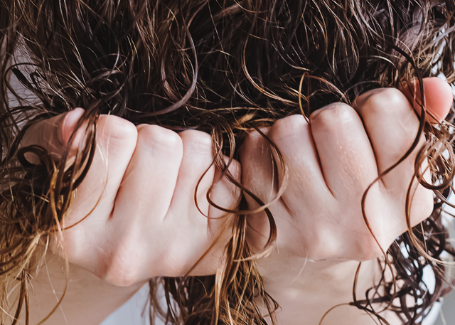 A fair-skinned woman scrunches her brown curly hair in front of a white background.