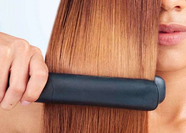 Woman with tan skin and yellow brown hair uses flat iron to straighten her hair in front of a grey background