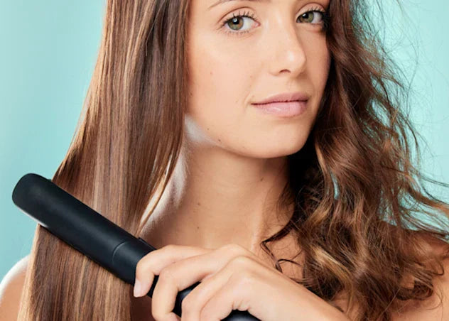 woman with fair skin and dark brown curly hair straightens it using a flat iron in front of a teal background.