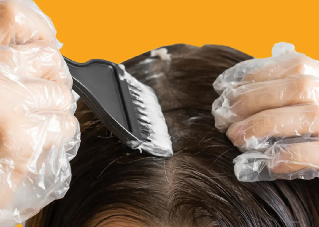 Woman with translucent gloves spreads bleach through her drak brown wet hair at the roots in front of a yellow background.