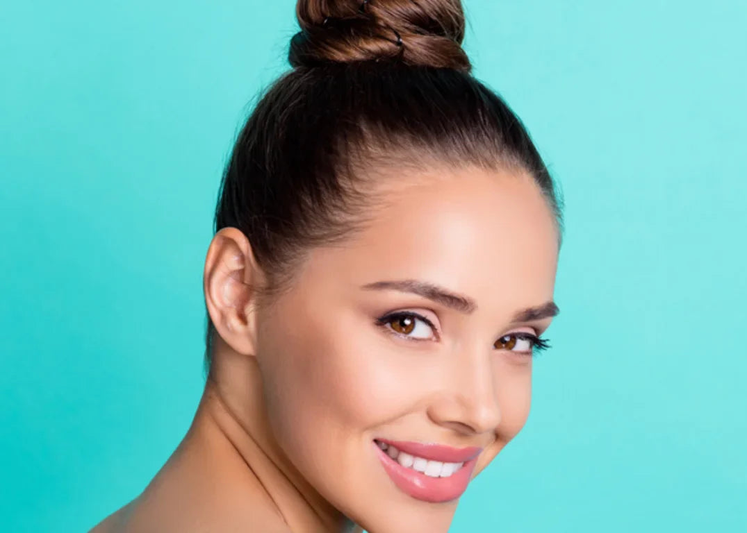 a woman with fair skin and a slicked back hair bun smiles and with a turned head facing the camera in front of a turquoise background.