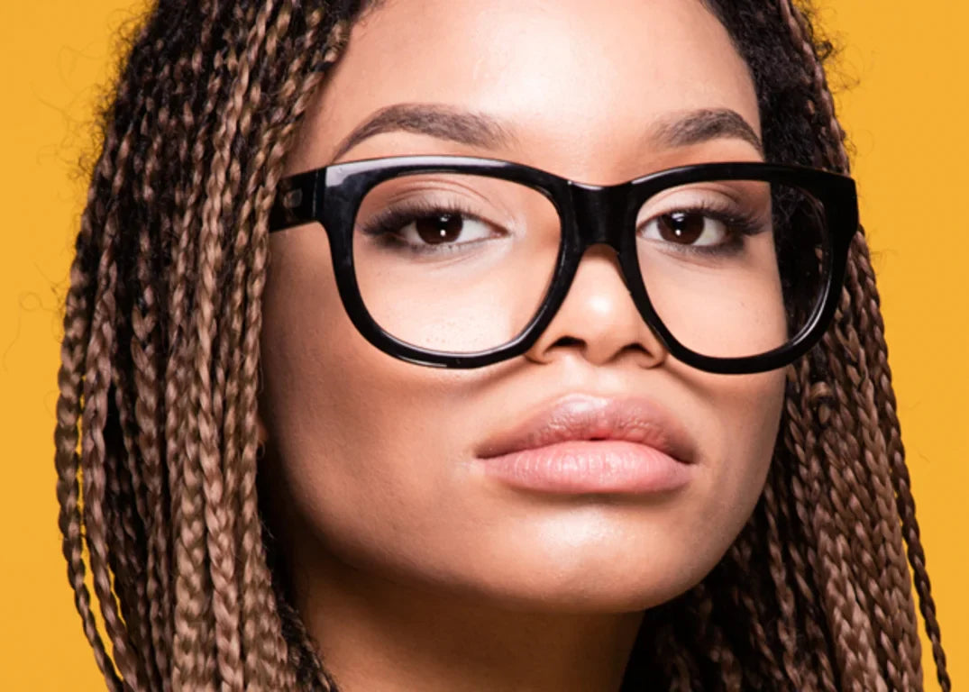 Dark skinned woman with black glasses and braided hair looks straight in front of a yellow background.