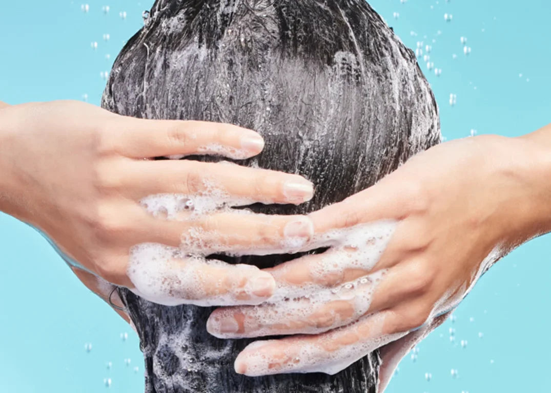 Woman with long dark hair and fair skin uses her hands to clean and wash her hair with bubbling shampoo after coloring it in front of a light blue background.