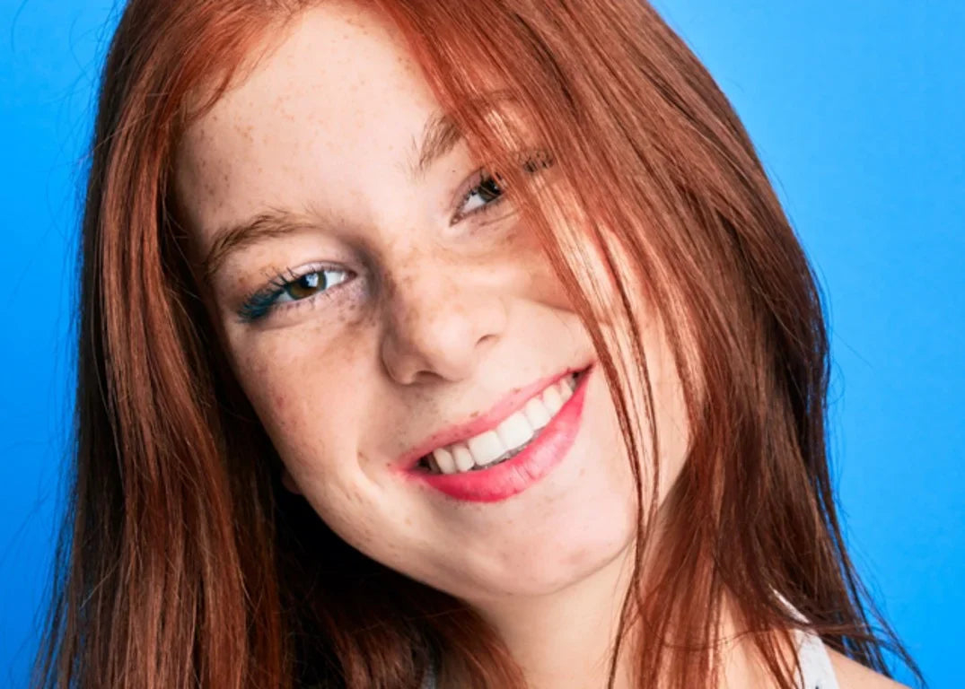 Woman with fair skin and freckles smiles with orange brown hair in front of a blue background.