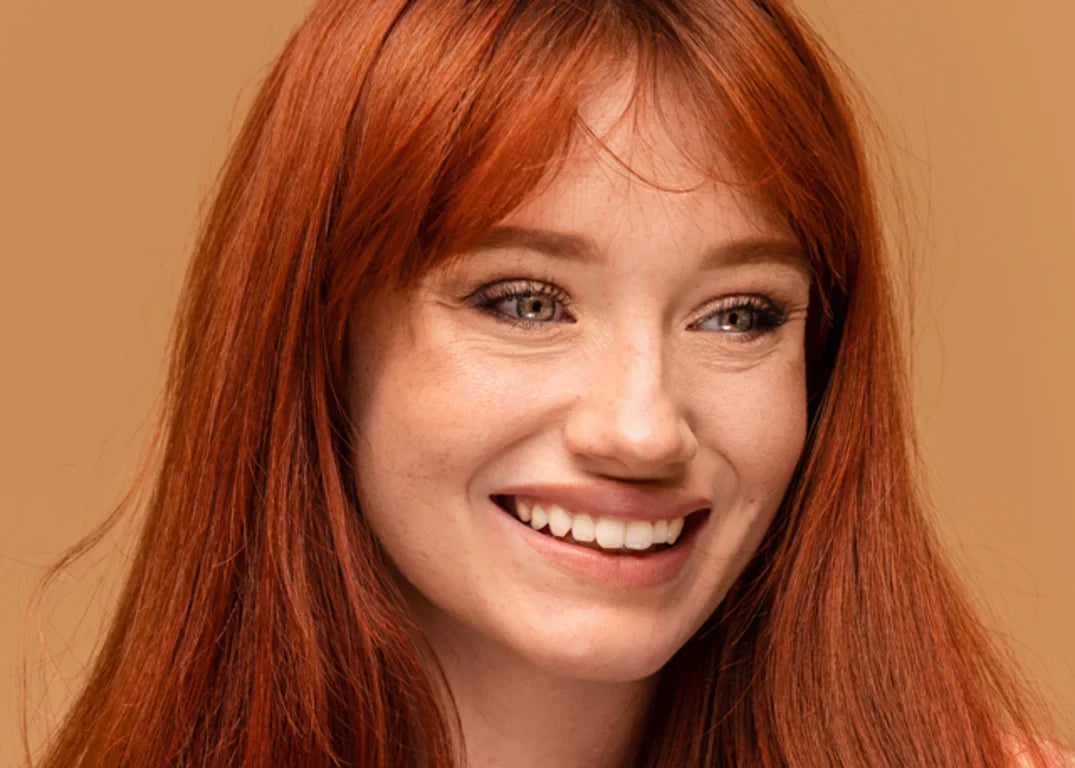Fair skinned woman with red colored hair smiles in front of an orange background.