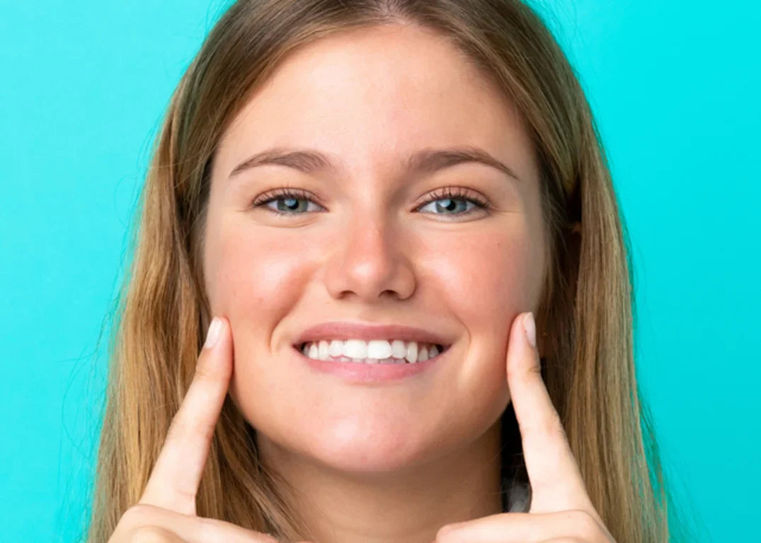 a woman with fair skin, green eyes, wavy blonde hair and a heart shaped face smiles forward in front of a light blue background.