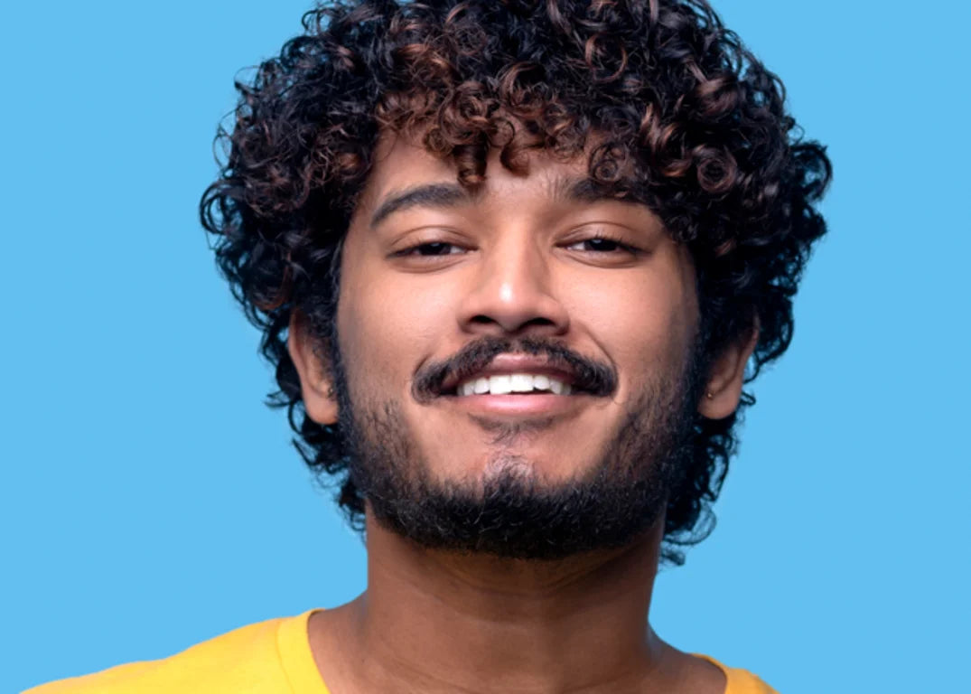 Man with curly brown hair and tan skin smiles facing forward in front of a blue background.