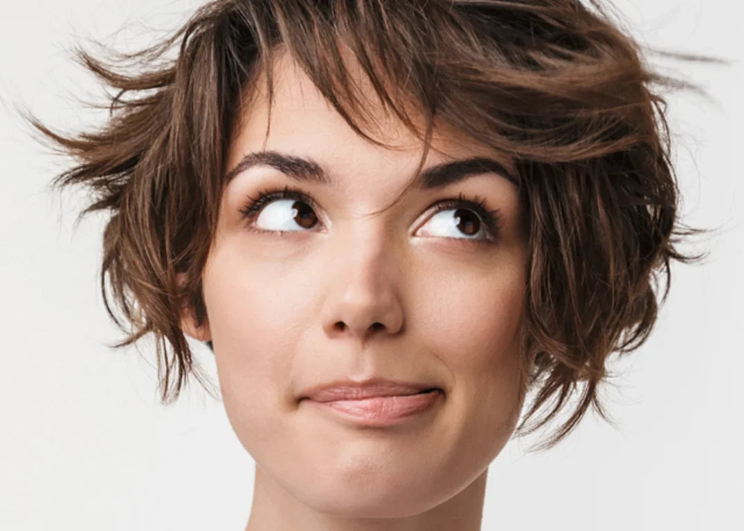 A woman with fair skin and choppy brown hair looks up to the left in front of a white background.