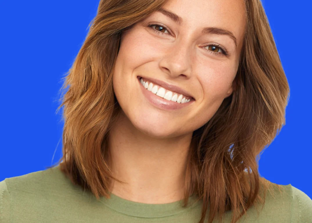 A woman with fair skin and medium length light brown smiles facing forward in front of a blue background.