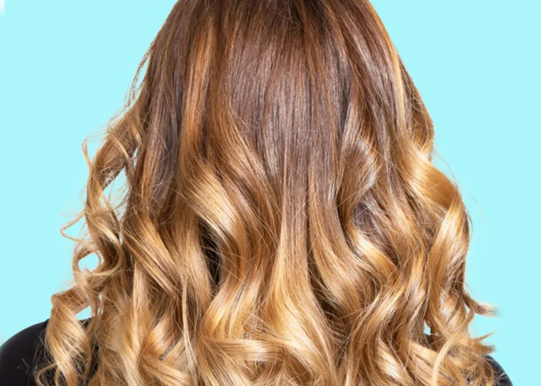 A person with a brown and blonde balayage hairstyle faces away in front of a light blue background.