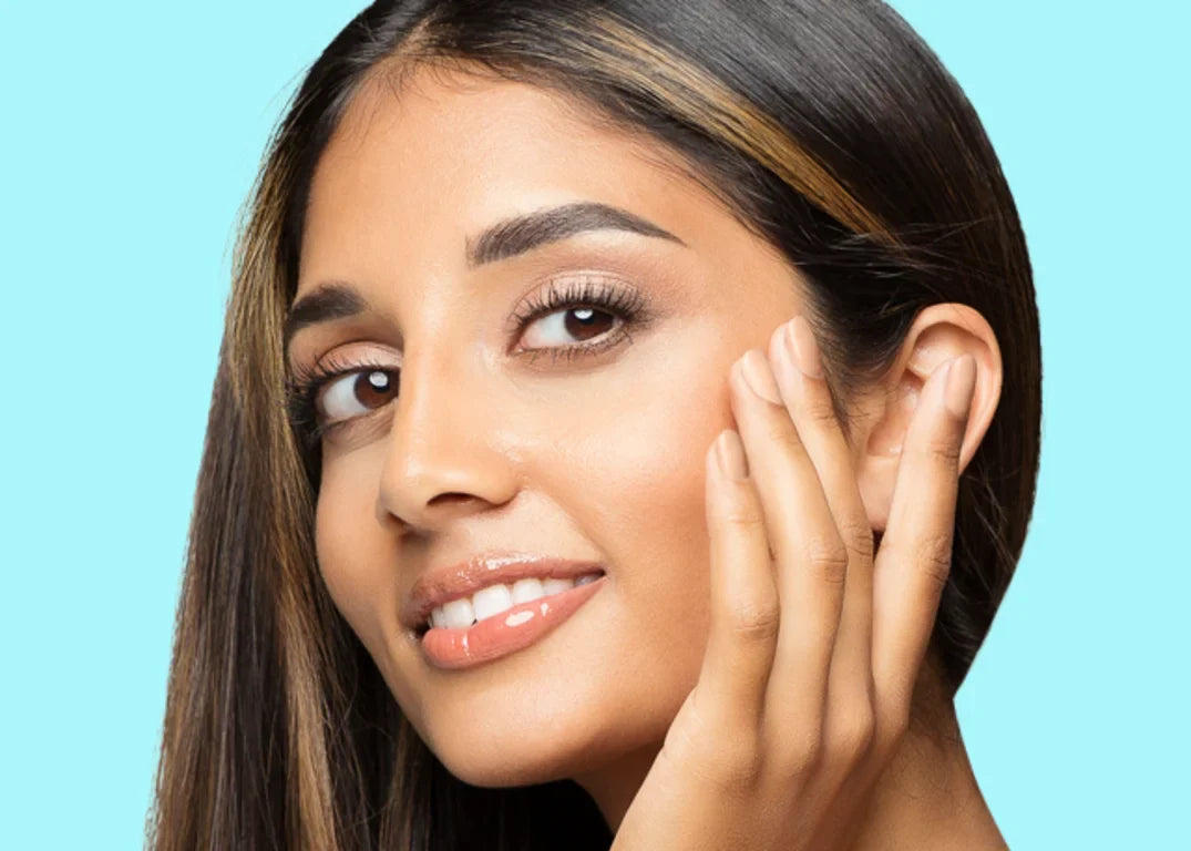 A woman with tan skin and highlights in her dark brown hair touches her face and looks forward.