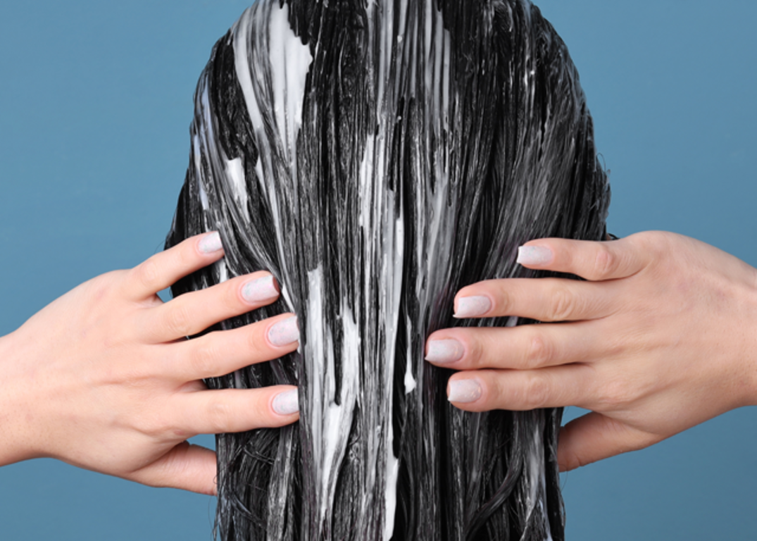 Fair skinned woman’s hands touch her black hair coated with hair conditioner in front of a blue background