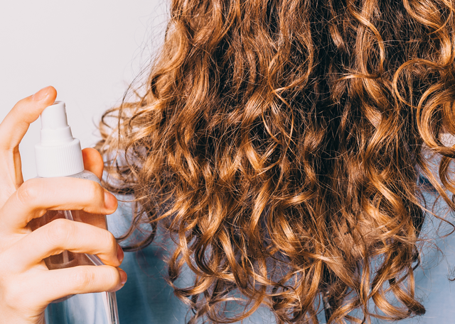 A fair-skinned woman holds a spray to refresh her brown curls in front of a white background.
