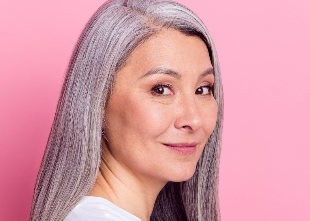 a woman with a round face and long gray hair turns her head to the right in front of a pink background.