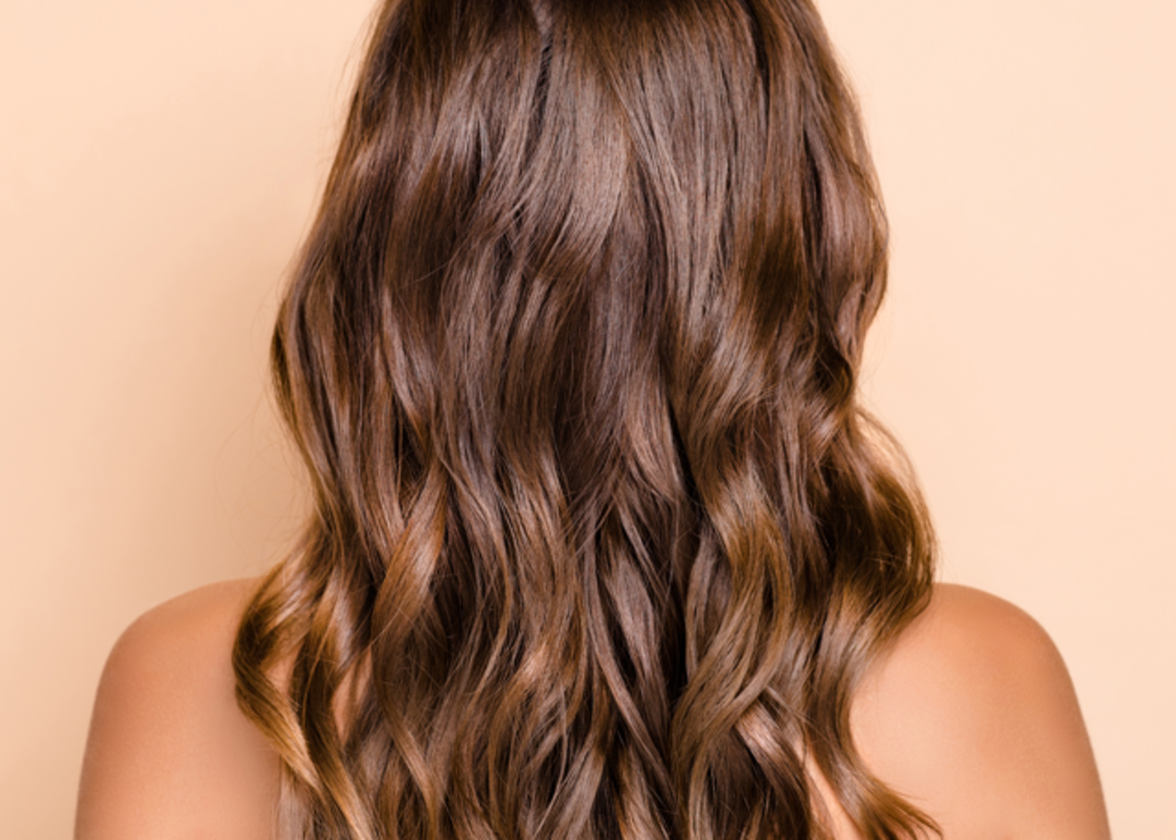 A woman with brown heat protected hair faces backwards in front of a light brown background.
