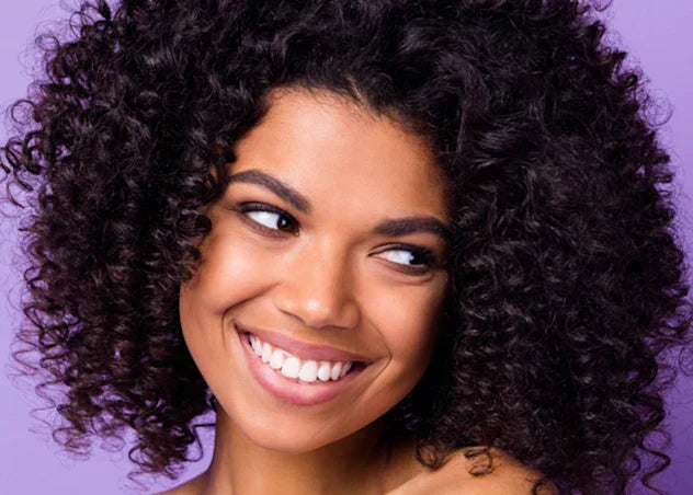 Tan skinned woman with long coily dark hair smiles in front of a purple background.