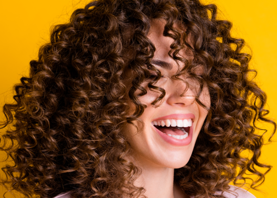 a woman with curly brown hair smiles with her eyes closed in front of a yellow background.