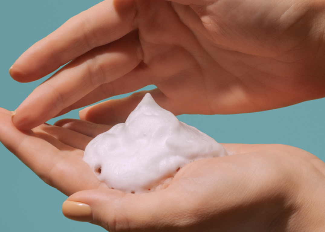 a person with fair skin holds mousse in the palm of their left hand in front of a gray blue background.