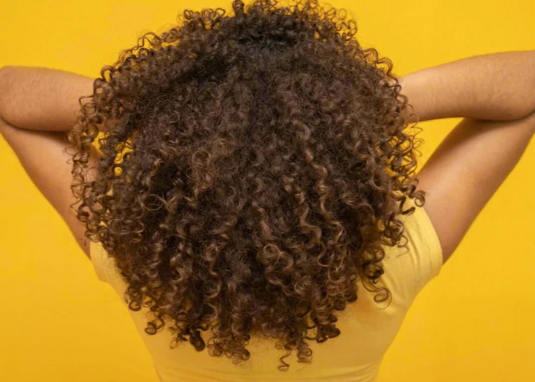 Dark-skinned woman holding coily brown hair in front of yellow background showing how to strengthen hair scalp to tip.