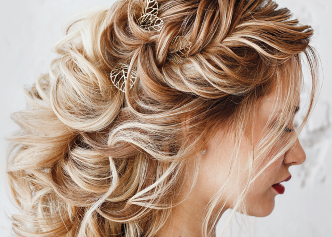 A woman with fair skin, wavy blonde hair in a prom hairstyle with leaf shaped clips looks down and right in front of a white background.