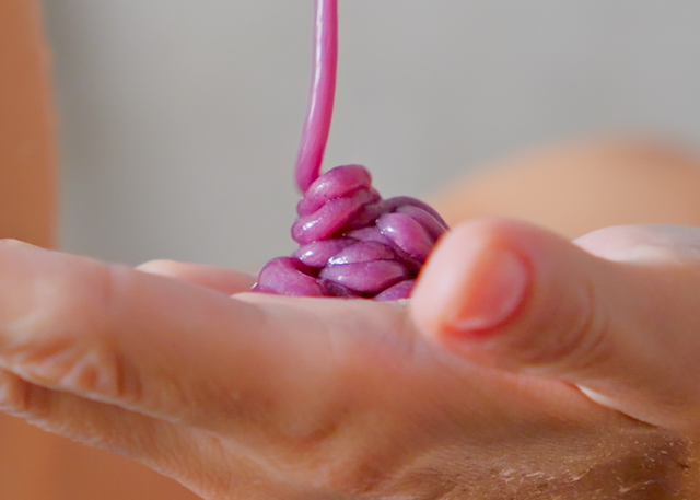 Purple shampoo is squeezed on a tan-skinned hand in front of a grey background.