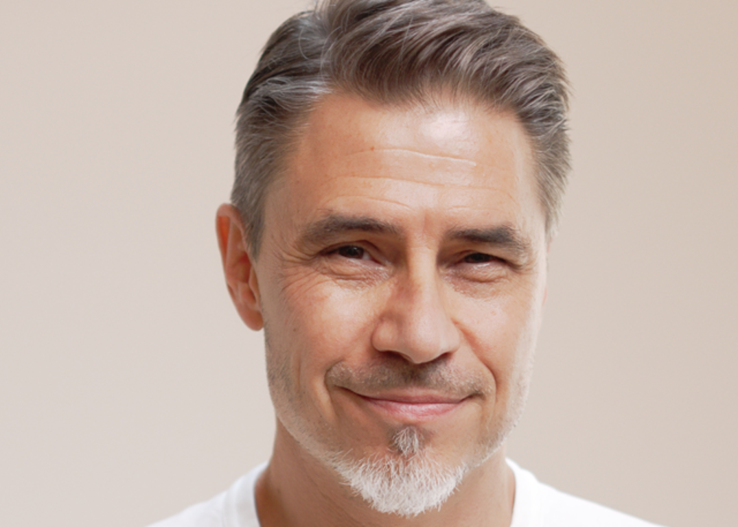 Senior man with fair skin and short gray hair smiles in front of a light brown background.