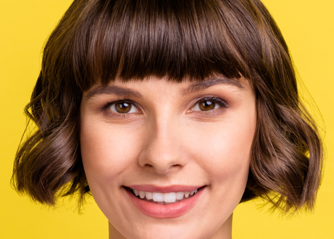 a woman with fair skin and a short brown bob hairstyle with bangs looks forward and smiles in front of a yellow background