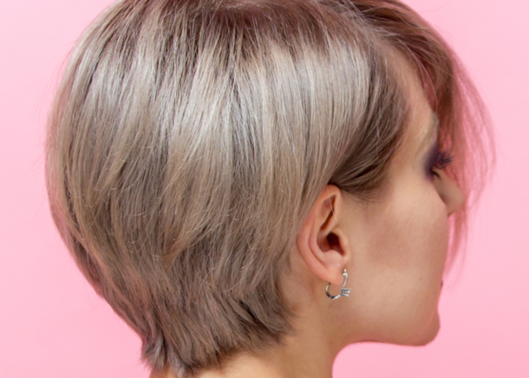 Woman with fair skin and grey-brown hair in Pixie hairstyle in front of a pink background.