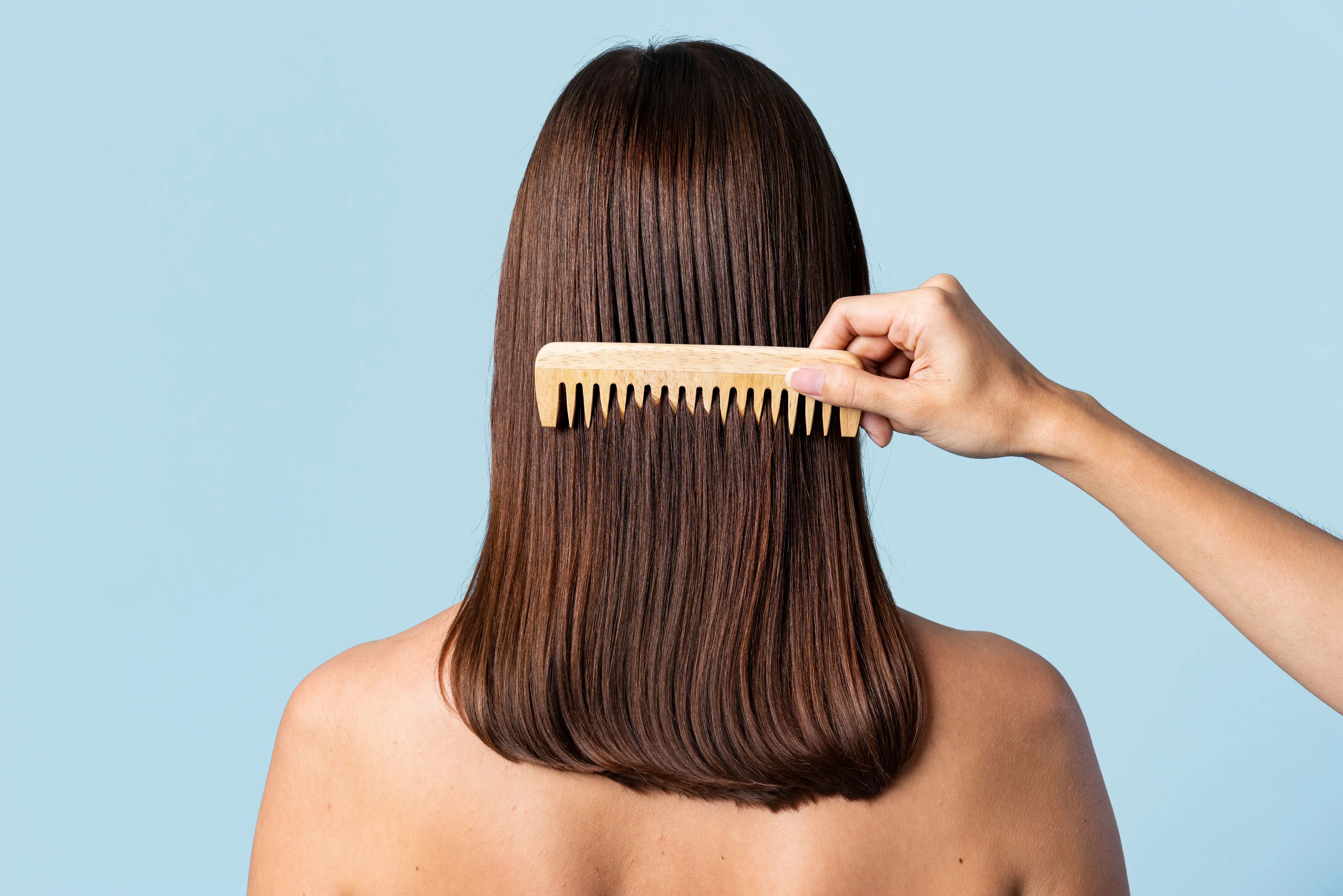 A person with long, straight brown hair has their hair combed against a light blue background after completing their personalized haircare routine. 