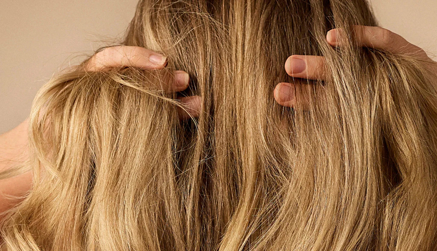 A woman using both hands to part blonde hair at the scalp, showing natural hair texture and roots
