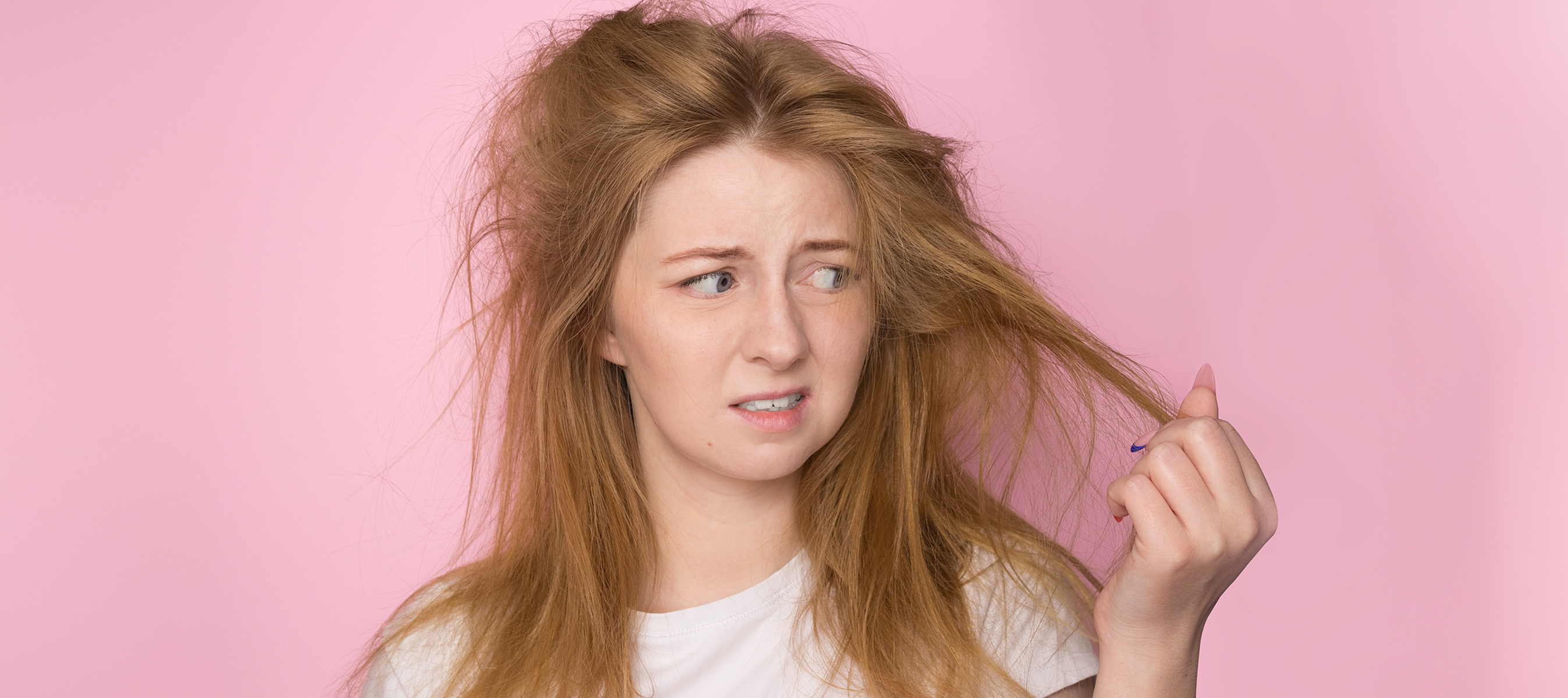 lifeless sun damaged hair. A woman on a pink background holds her disheveled, tangled hair and looks at it with a discontented expression.