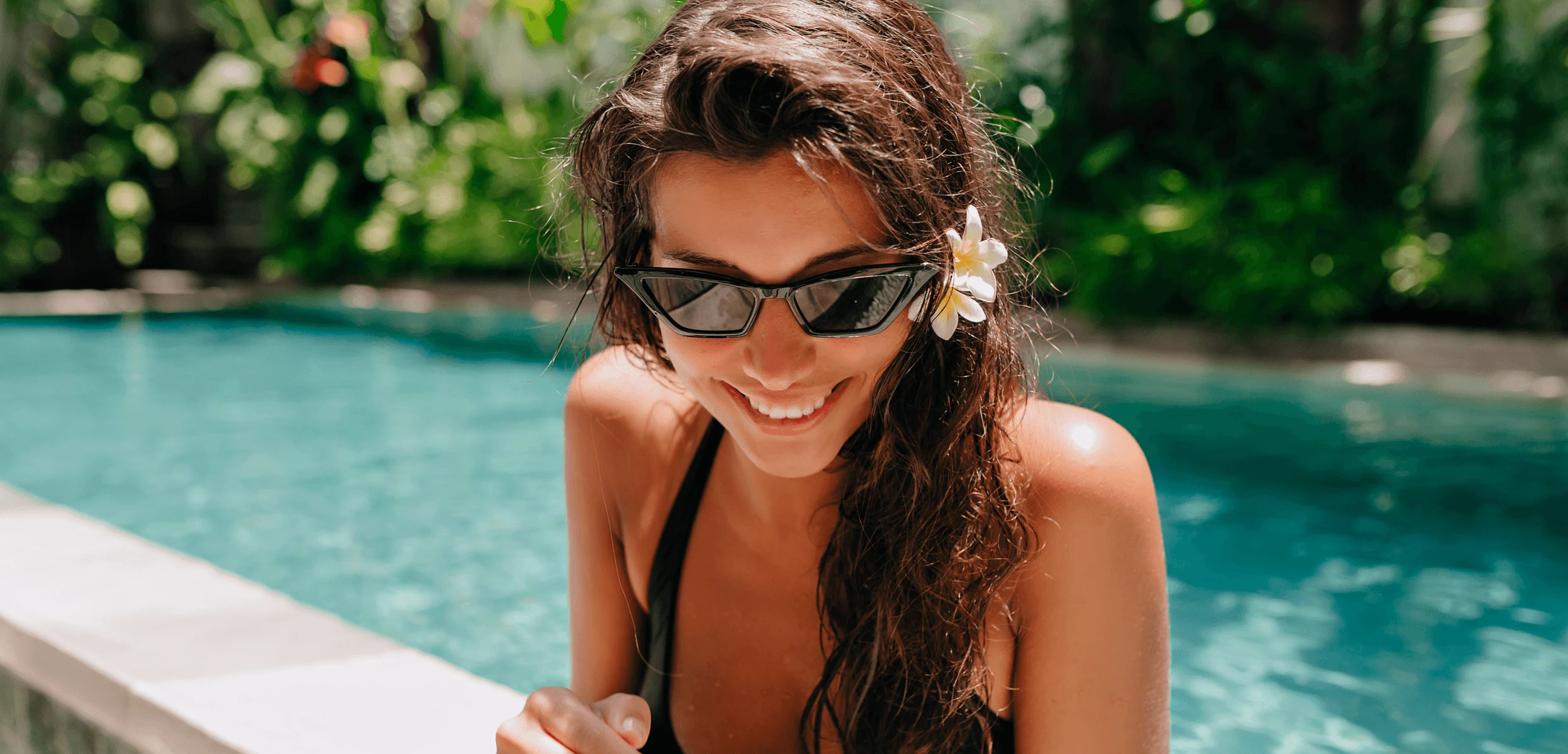 Smiling brunette woman with sunglasses and daisy in hair leaning on edge of pool